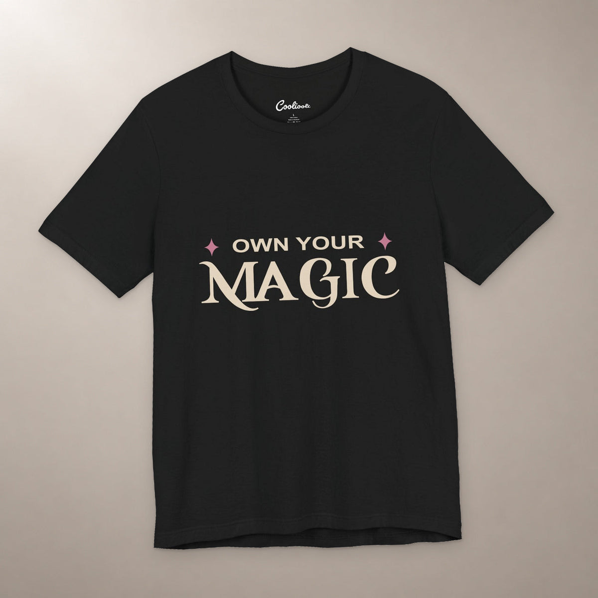 She's Got That Something. Bella+Canvas 3001 T-shirt in Black, front flat lay with Own Your Magic lettering and two rose-coloured stars on a plain warm neutral background.