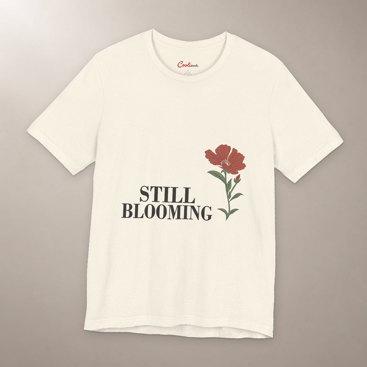 Still Coming Into Her Own. Bella+Canvas 3001 T-shirt in Natural, front flat lay with Still Blooming lettering and a brick-red poppy on a plain warm neutral background.