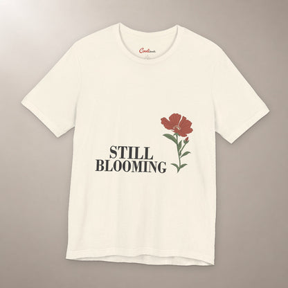 Still Coming Into Her Own. Bella+Canvas 3001 T-shirt in Natural, front flat lay with Still Blooming lettering and a brick-red poppy on a plain warm neutral background.
