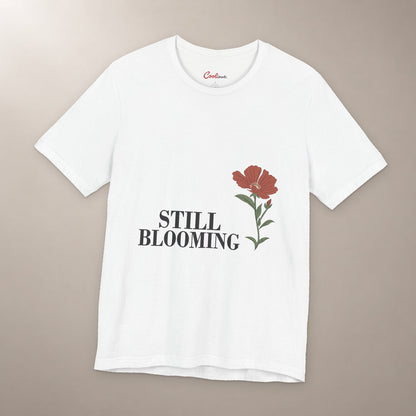 Still Coming Into Her Own. Bella+Canvas 3001 T-shirt in White, front flat lay with Still Blooming lettering and a brick-red poppy on a plain warm neutral background.