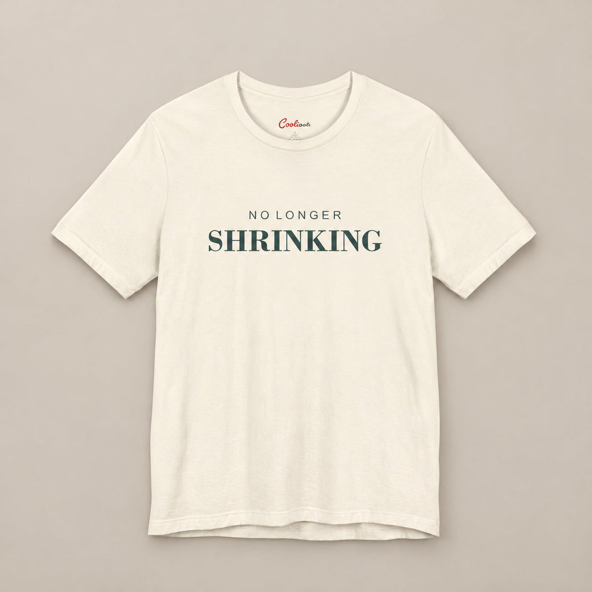 Natural NO LONGER SHRINKING Bella+Canvas 3001 tee, complete square flat lay with deep green lettering on a plain warm neutral background.