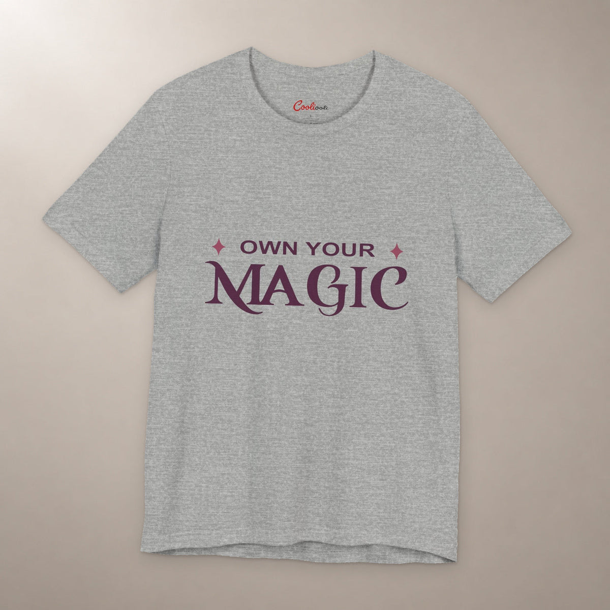 She's Got That Something. Bella+Canvas 3001 T-shirt in Athletic Heather, front flat lay with Own Your Magic lettering and two rose-coloured stars on a plain warm neutral background.