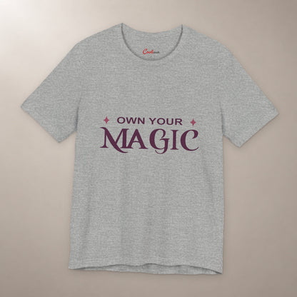 She's Got That Something. Bella+Canvas 3001 T-shirt in Athletic Heather, front flat lay with Own Your Magic lettering and two rose-coloured stars on a plain warm neutral background.