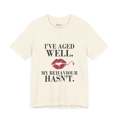 Natural Cooliooli Bella+Canvas 3001 T-shirt with “I’ve aged well. My behaviour hasn’t.” lettering and lipstick kiss with devil tail, front flat lay