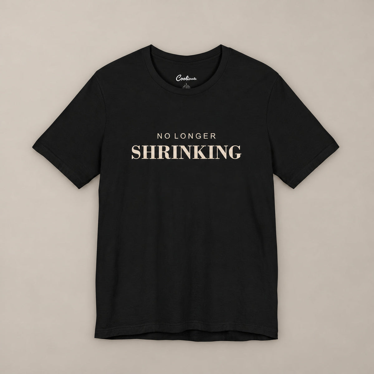 Black NO LONGER SHRINKING Bella+Canvas 3001 tee, complete square flat lay with warm ivory lettering on a plain warm neutral background.