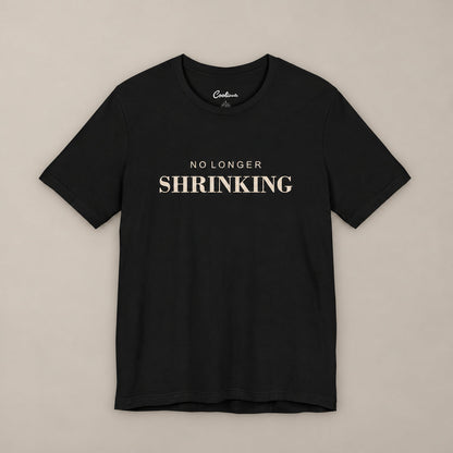 Black NO LONGER SHRINKING Bella+Canvas 3001 tee, complete square flat lay with warm ivory lettering on a plain warm neutral background.