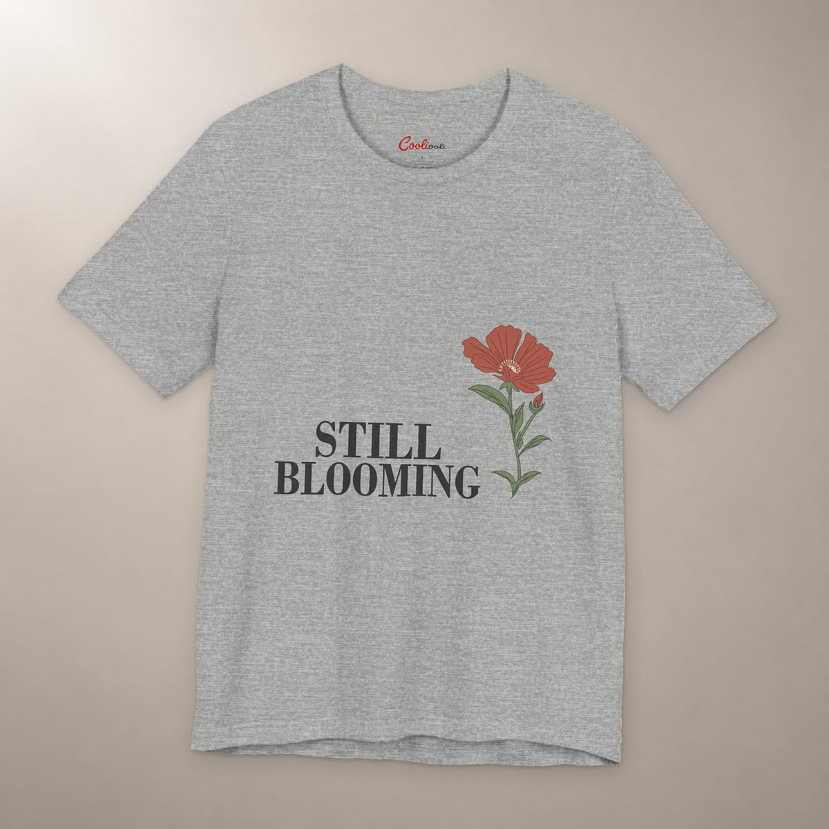 Still Coming Into Her Own. Bella+Canvas 3001 T-shirt in Athletic Heather, front flat lay with Still Blooming lettering and a brick-red poppy on a plain warm neutral background.