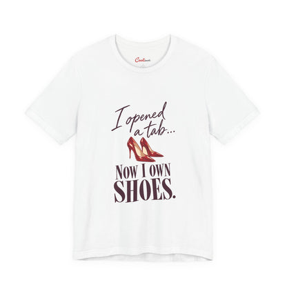 White Cooliooli T-shirt with red heels and “I opened a tab… Now I own shoes.” lettering, front flat lay