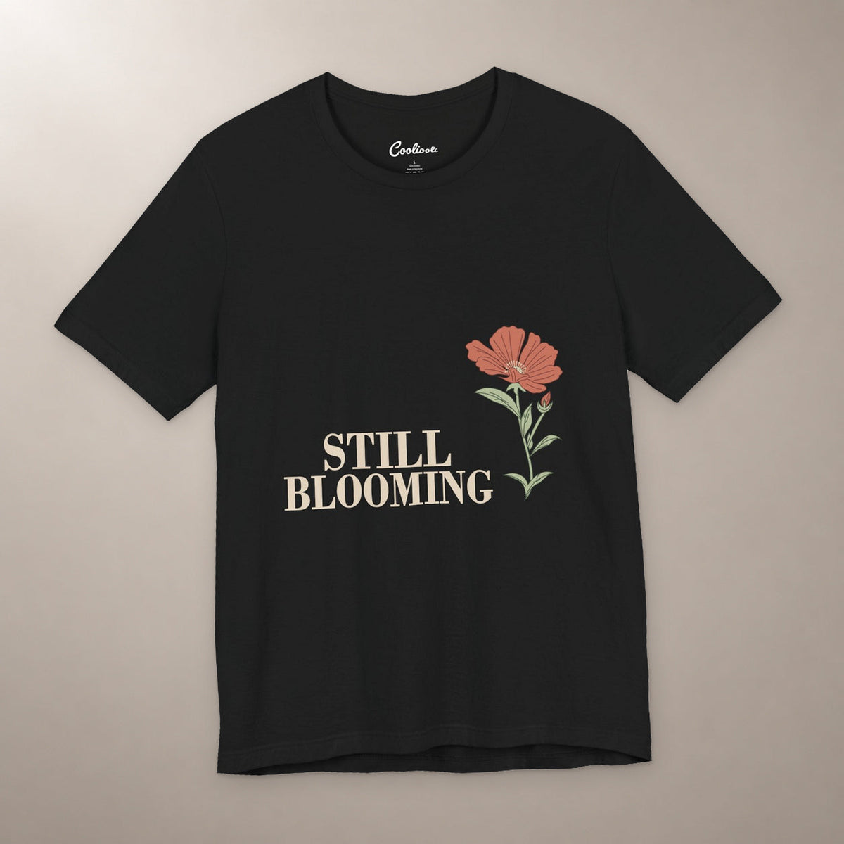 Still Coming Into Her Own. Bella+Canvas 3001 T-shirt in Black, front flat lay with Still Blooming lettering and a brick-red poppy on a plain warm neutral background.