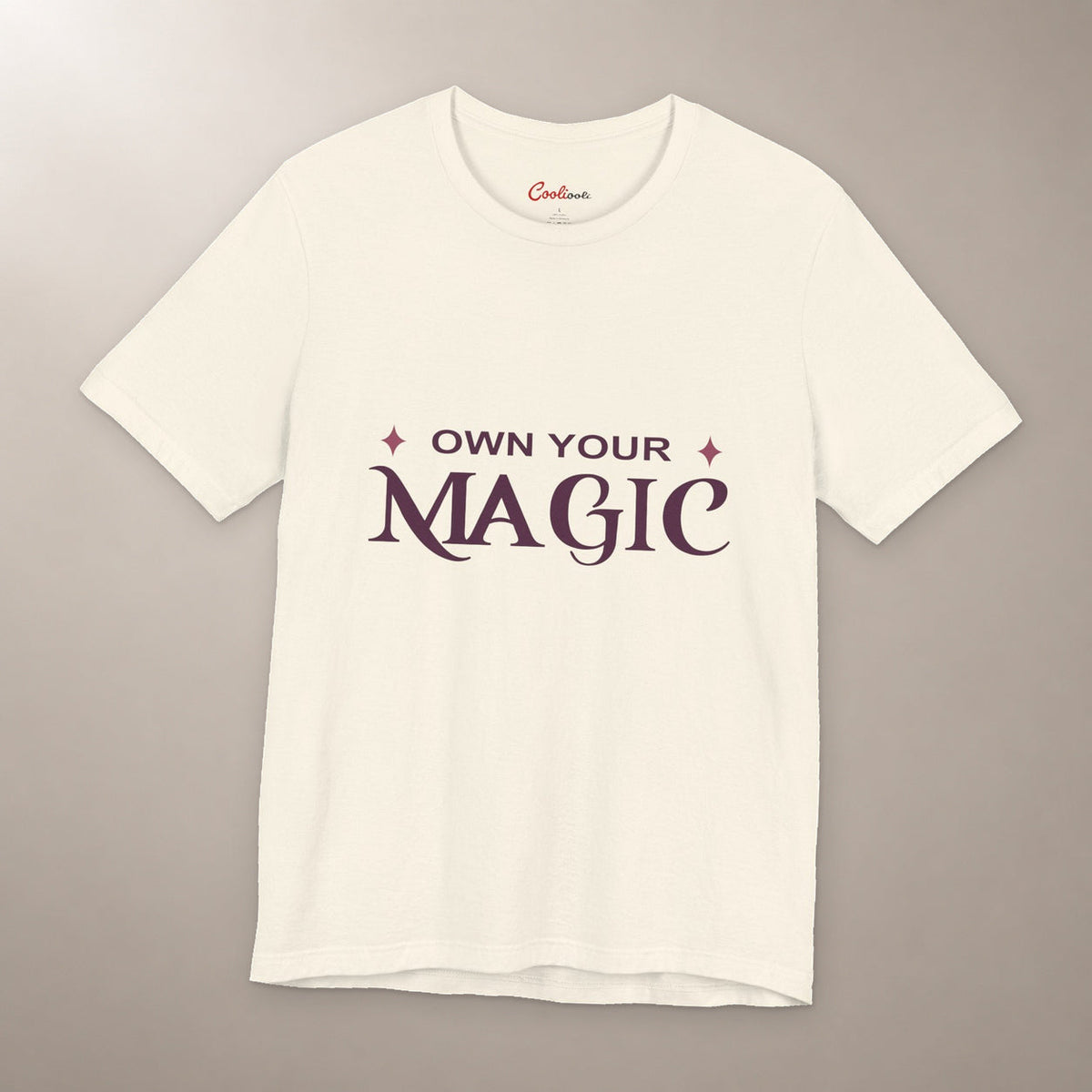She's Got That Something. Bella+Canvas 3001 T-shirt in Natural, front flat lay with Own Your Magic lettering and two rose-coloured stars on a plain warm neutral background.