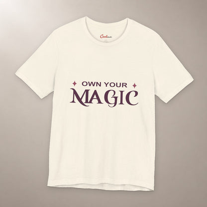 She's Got That Something. Bella+Canvas 3001 T-shirt in Natural, front flat lay with Own Your Magic lettering and two rose-coloured stars on a plain warm neutral background.