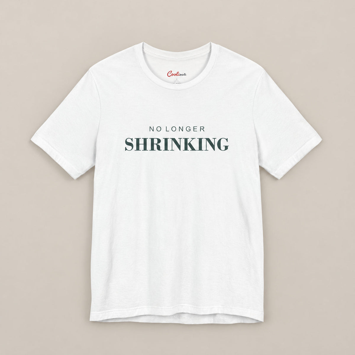 White NO LONGER SHRINKING Bella+Canvas 3001 tee, complete square flat lay with deep green lettering on a plain warm neutral background.