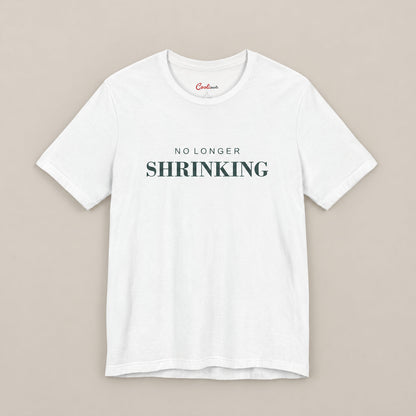 White NO LONGER SHRINKING Bella+Canvas 3001 tee, complete square flat lay with deep green lettering on a plain warm neutral background.
