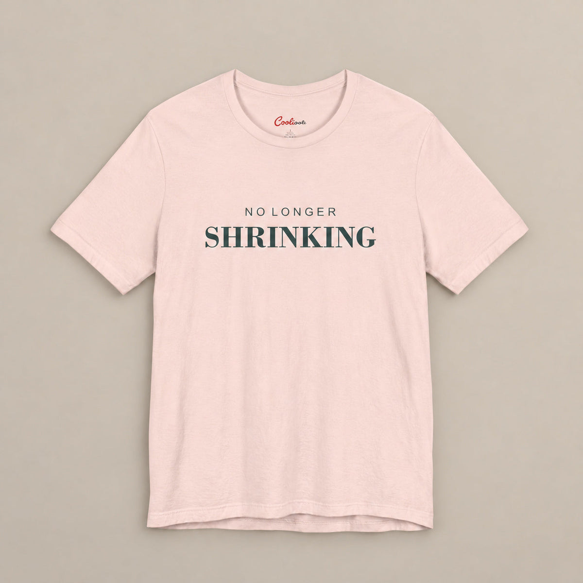 Soft Pink NO LONGER SHRINKING Bella+Canvas 3001 tee, complete square flat lay with deep green lettering on a plain warm neutral background.