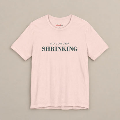 Soft Pink NO LONGER SHRINKING Bella+Canvas 3001 tee, complete square flat lay with deep green lettering on a plain warm neutral background.