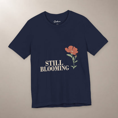 Still Coming Into Her Own. Bella+Canvas 3001 T-shirt in Navy, front flat lay with Still Blooming lettering and a brick-red poppy on a plain warm neutral background.