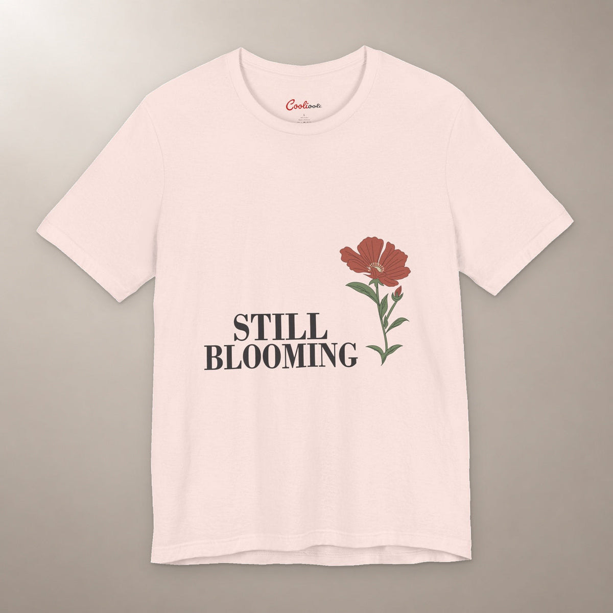 Still Coming Into Her Own. Bella+Canvas 3001 T-shirt in Soft Pink, front flat lay with Still Blooming lettering and a brick-red poppy on a plain warm neutral background.
