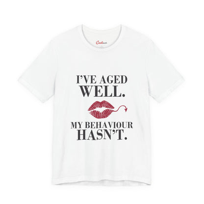 White Cooliooli Bella+Canvas 3001 T-shirt with “I’ve aged well. My behaviour hasn’t.” lettering and lipstick kiss with devil tail, front flat lay