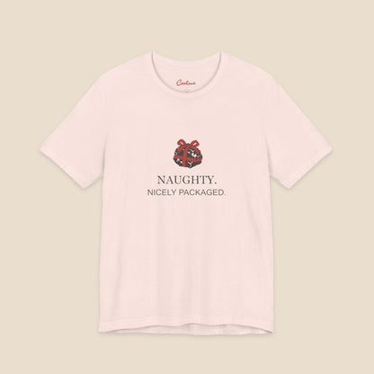 Soft Pink Bella+Canvas 3001 Christmas T-shirt with an original coal and red bow illustration above Naughty. Nicely Packaged. lettering.