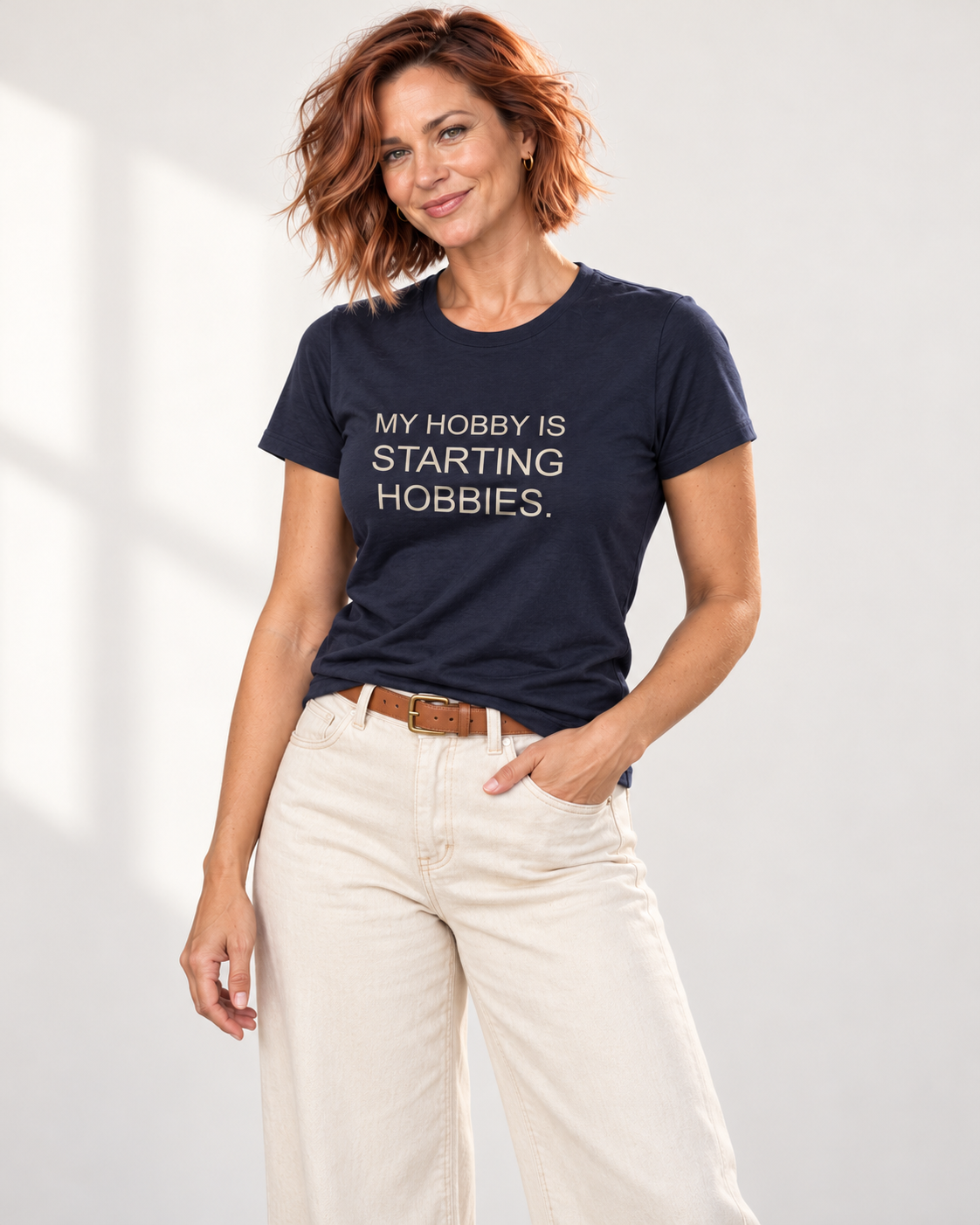 Sienna wearing the Navy My Hobby Is Starting Hobbies Bella+Canvas3001 tee with ecru denim and a tan belt in the pale studio.