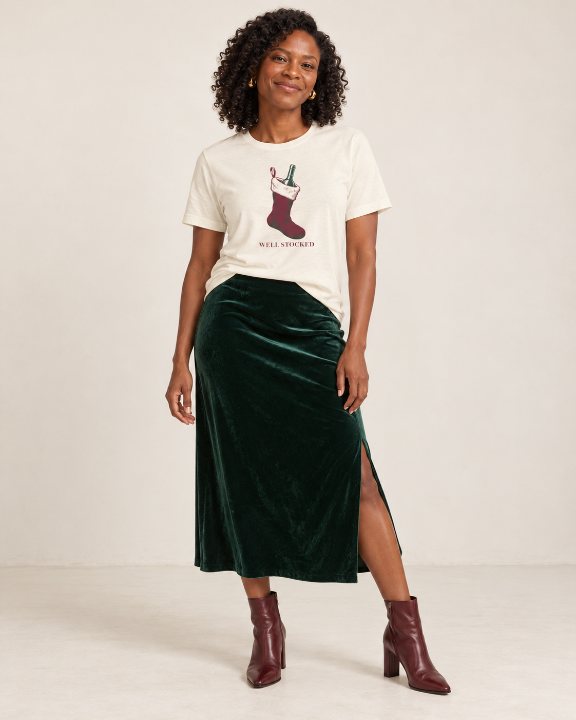 Amara wearing the Natural Well Stocked Christmas wine-stocking T-shirt with a forest-green velvet midi skirt and oxblood ankle boots.