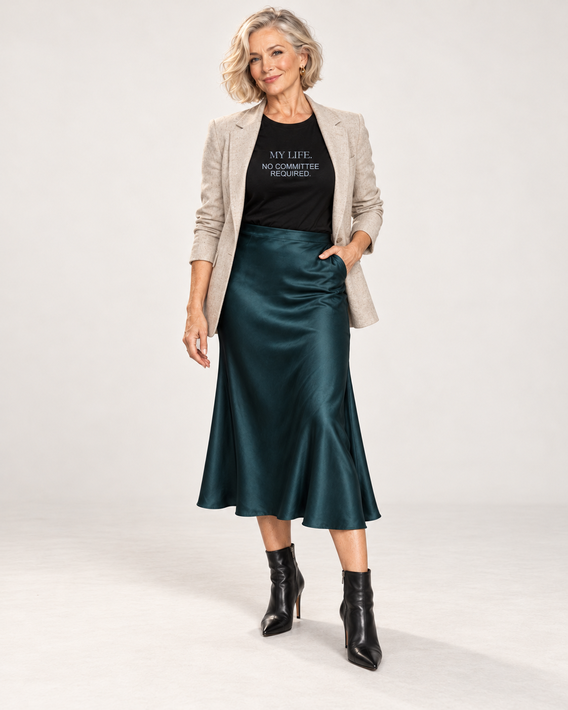 Claudia wearing the Black My Life No Committee Required Bella+Canvas3001 unisex tee with an open oatmeal blazer, deep teal satin midi skirt and pointed black ankle boots in a pale studio.