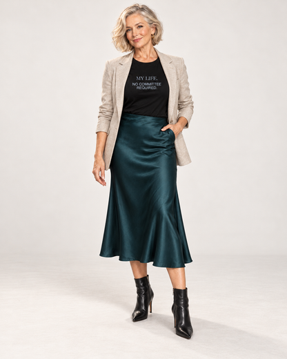 Claudia wearing the Black My Life No Committee Required Bella+Canvas3001 unisex tee with an open oatmeal blazer, deep teal satin midi skirt and pointed black ankle boots in a pale studio.