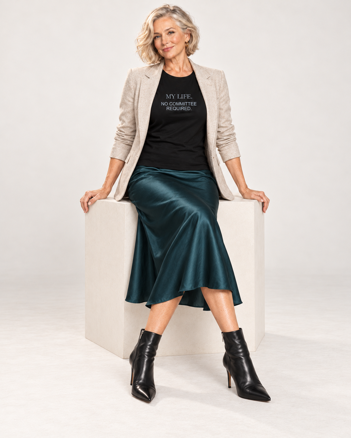 Claudia wearing the Black My Life No Committee Required Bella+Canvas6400 women’s relaxed-fit tee with an open oatmeal blazer, deep teal satin midi skirt and pointed black ankle boots, seated in a pale studio.