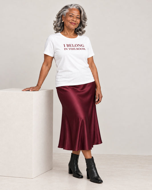 Vivienne wearing the White Bella+Canvas3001 T-shirt with deep burgundy satin midi skirt with black heeled ankle boots. Exact print: I BELONG IN THIS ROOM.