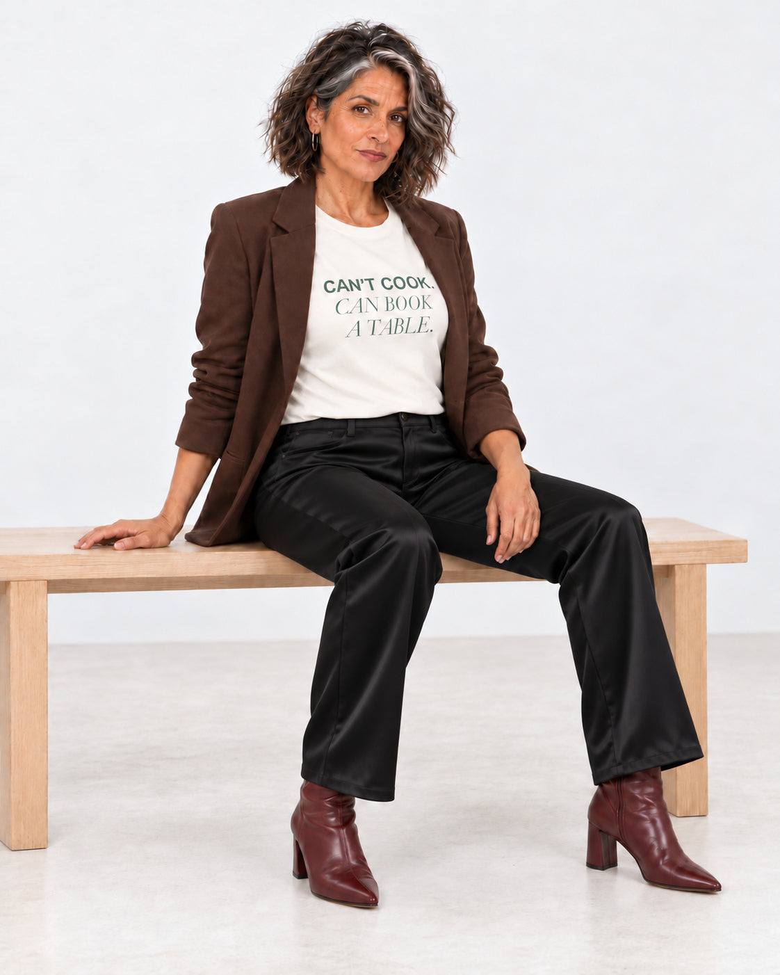 Freya wearing the Natural Can't Cook, Can Book a Table Bella+Canvas 3001 tee with an open blazer, black satin trousers and heeled boots.