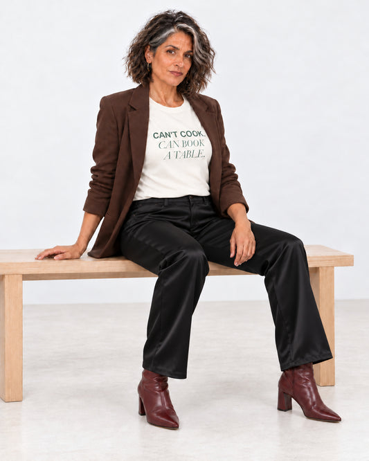 Freya wearing the Natural Can't Cook, Can Book a Table Bella+Canvas 3001 tee with an open blazer, black satin trousers and heeled boots.