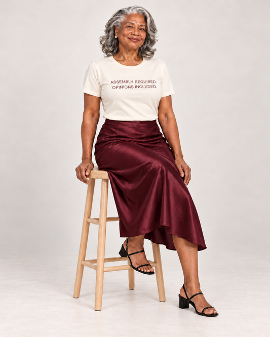 Vivienne wearing the Natural Assembly Required. Opinions Included. T-shirt with a burgundy satin midi skirt and strappy heels, seated on a wooden stool.