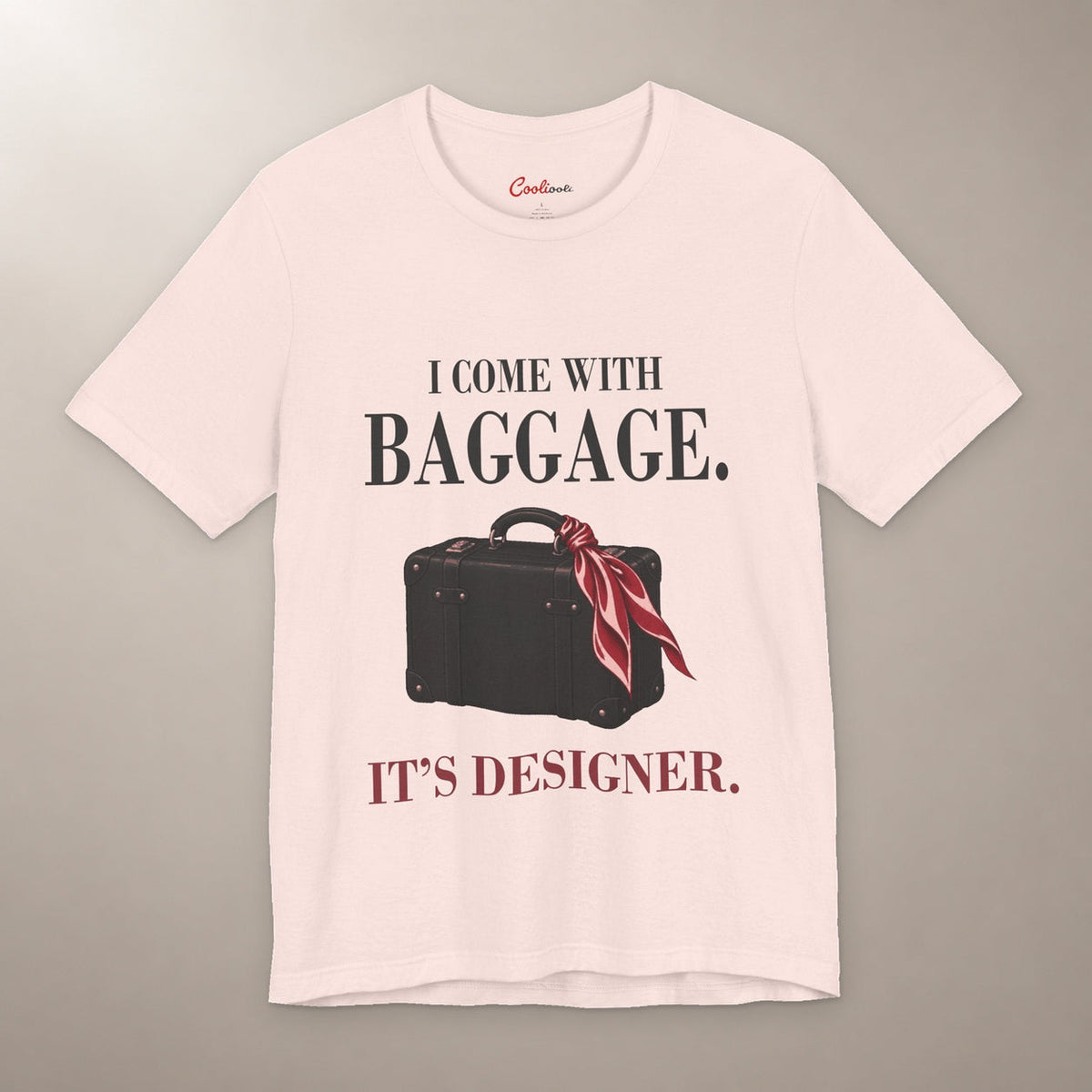 Excess Baggage. Excellent Taste. Bella+Canvas 3001 T-shirt in Soft Pink, front flat lay with I come with baggage. It’s designer. lettering and a vintage suitcase with a burgundy scarf on a plain warm neutral background.