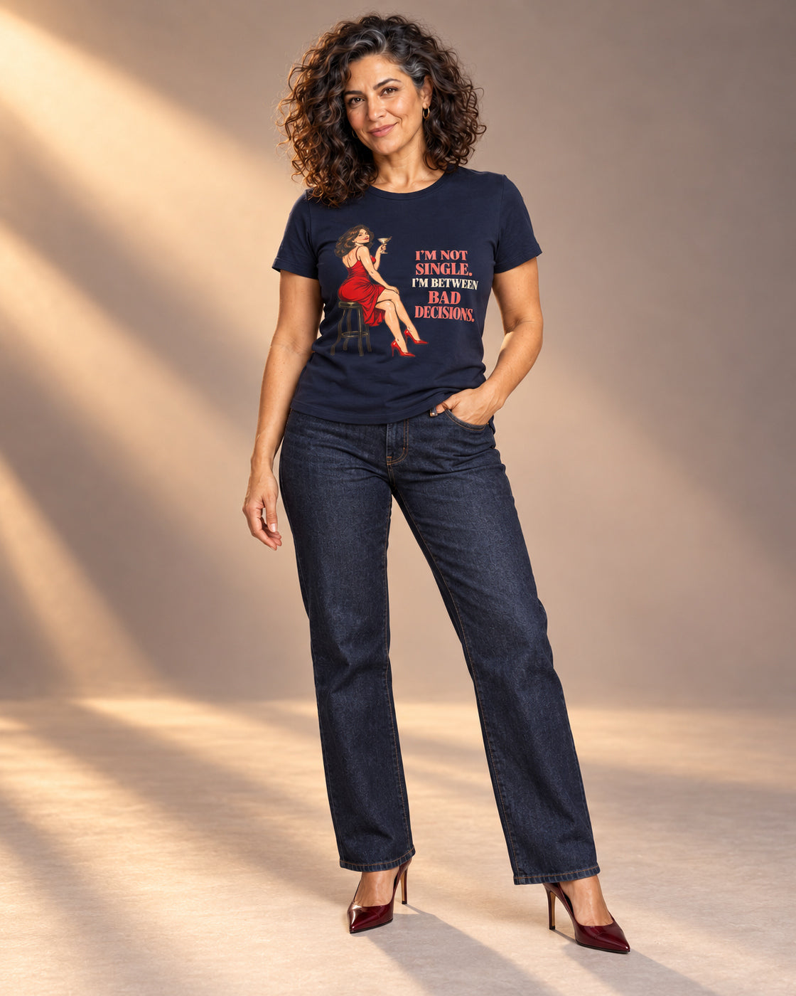 Leila models the Navy Bella+Canvas 3001 Between Bad Decisions tee with dark jeans and wine-red heels in a professional studio photoshoot.