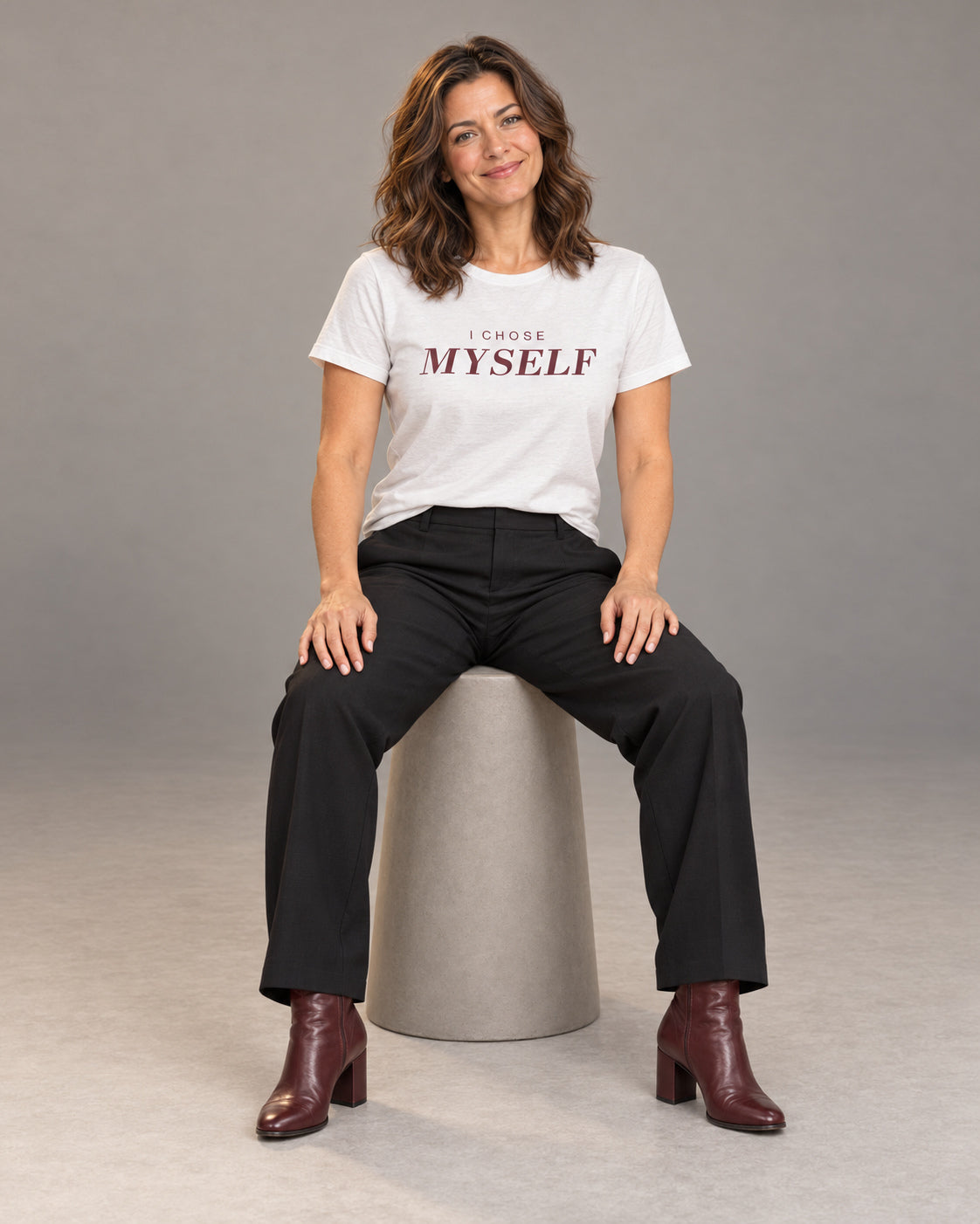 Sophie wearing the White I CHOSE MYSELF Bella+Canvas 3001 tee with black straight-leg trousers and burgundy heeled ankle boots, seated full-length studio portrait.