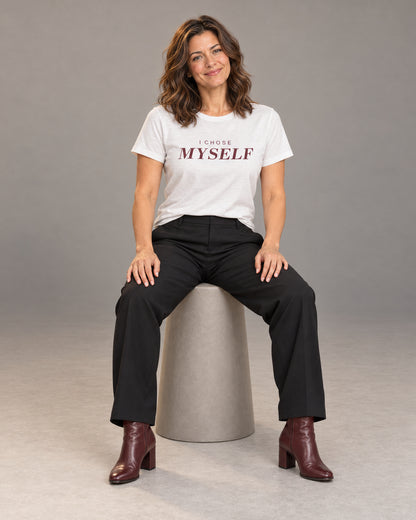 Sophie wearing the White I CHOSE MYSELF Bella+Canvas 3001 tee with black straight-leg trousers and burgundy heeled ankle boots, seated full-length studio portrait.