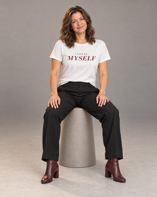 Sophie wearing the White I CHOSE MYSELF Bella+Canvas 3001 tee with black straight-leg trousers and burgundy heeled ankle boots, seated full-length studio portrait.