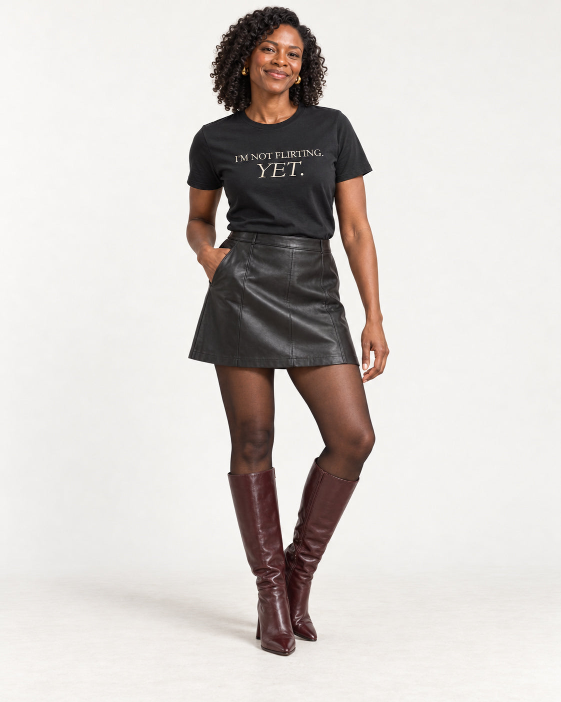Amara wearing the black I'm Not Flirting Yet T-shirt with a leather-look mini and burgundy heeled boots