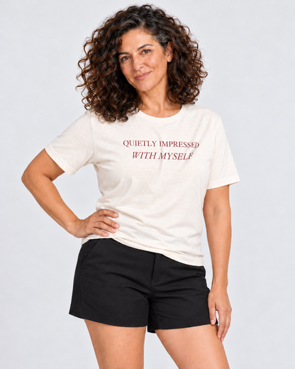 Leila wearing the Natural Quietly Impressed With Myself T-shirt and tailored black shorts in a pale studio, hand resting on her hip.