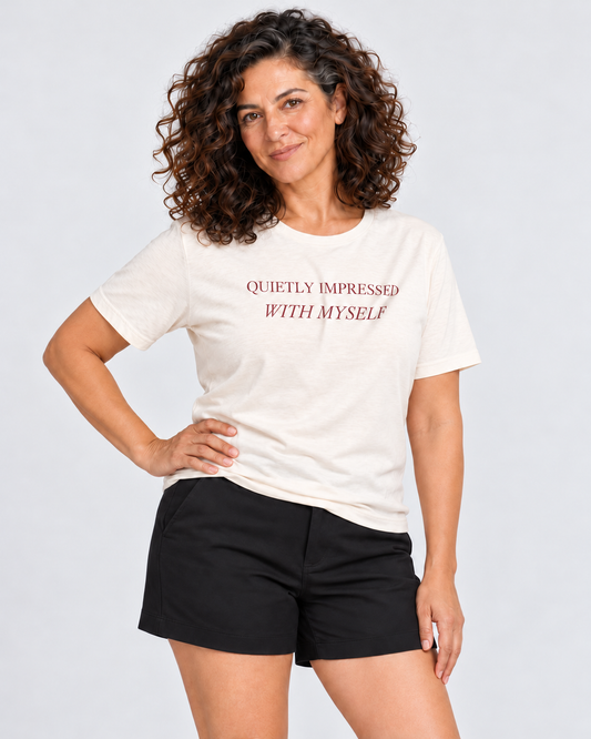 Leila wearing the Natural Quietly Impressed With Myself T-shirt and tailored black shorts in a pale studio, hand resting on her hip.