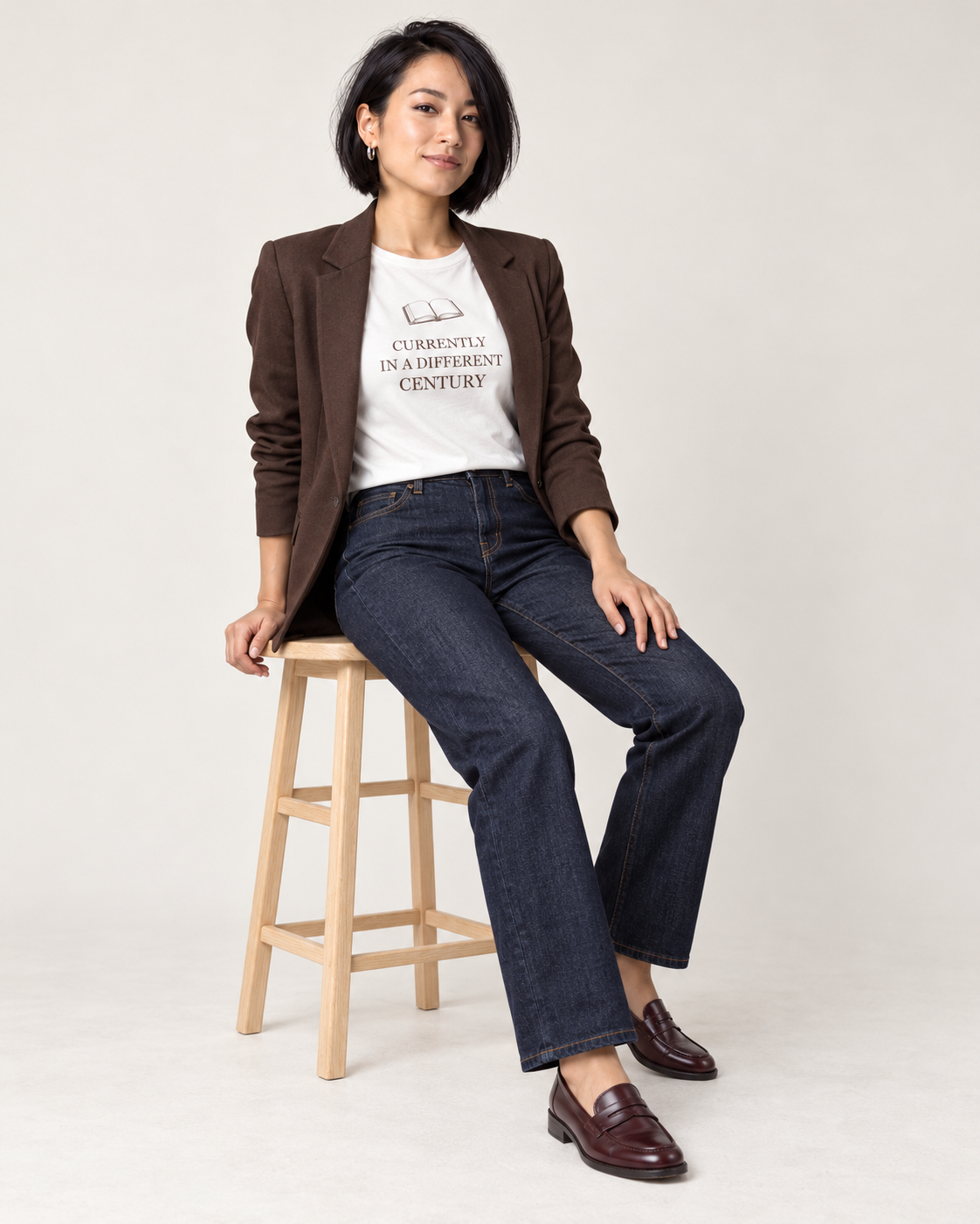 Mina wearing the White historical-fiction reader T-shirt with an open chocolate blazer, dark straight-leg jeans and oxblood loafers, seated on a wooden stool.