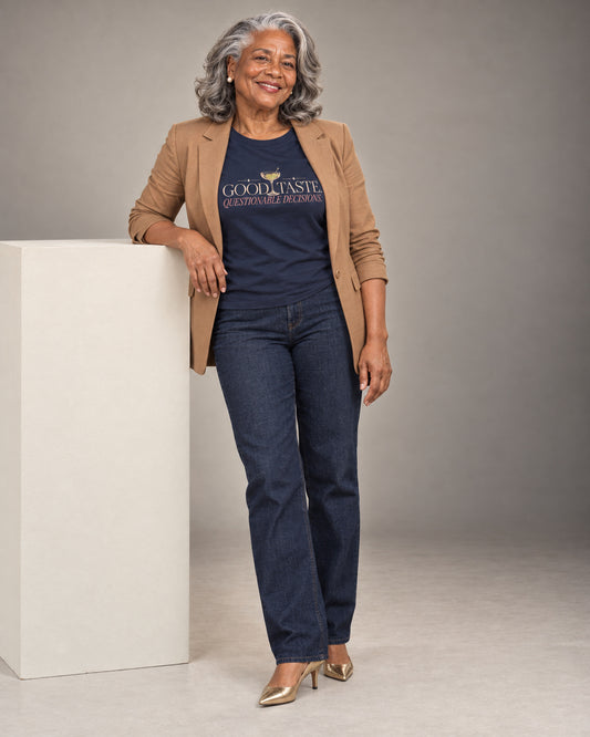 Vivienne leans beside a cream studio plinth wearing the Navy Good taste. Questionable decisions. Cooliooli tee with a caramel blazer, dark jeans and gold heels, smiling warmly in soft studio light.