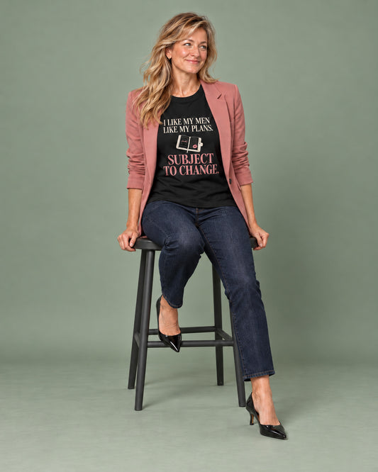 Elena sits casually on a studio stool wearing the Black Subject to change Cooliooli tee with a rose blazer, dark jeans and black heels, smiling towards someone off-camera.