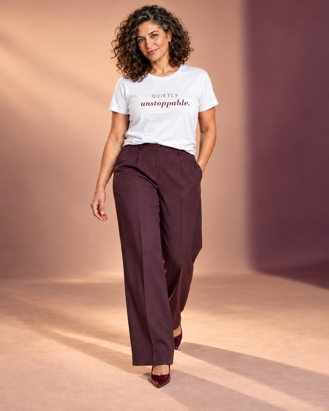 Leila wearing the white Quietly Unstoppable tee with plum tailored trousers and burgundy heels, walking in an apricot and plum studio.