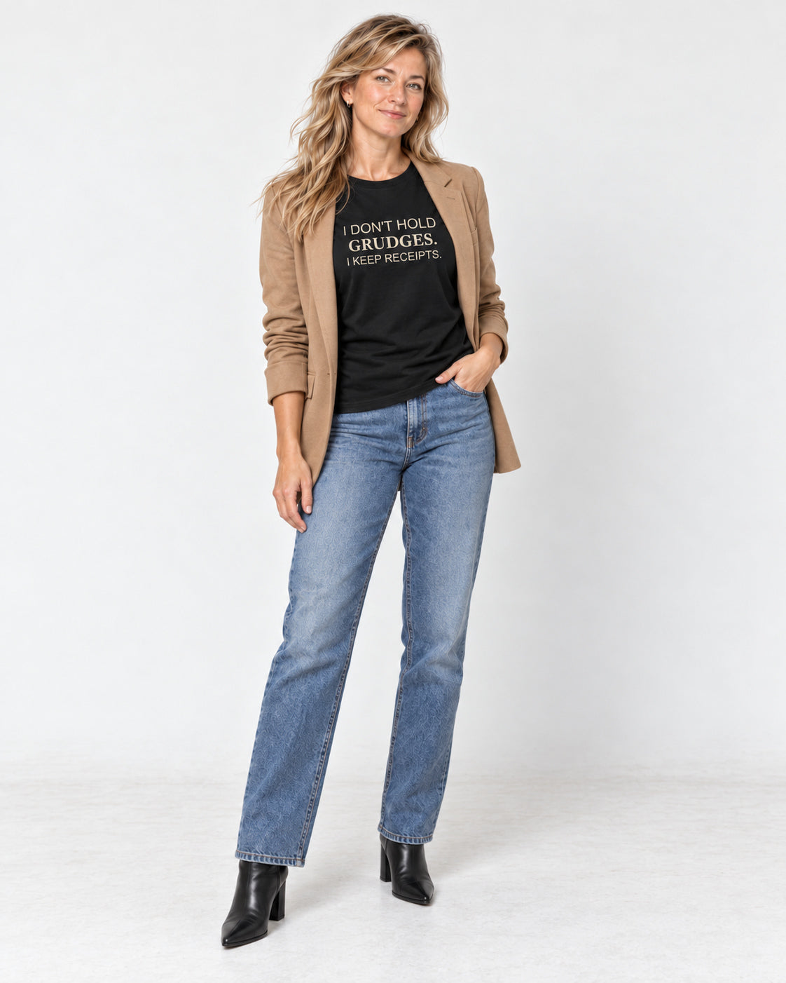 Elena wearing the Black I Don't Hold Grudges. I Keep Receipts. Bella+Canvas3001 T-shirt with an open camel blazer, straight-leg jeans and pointed black ankle boots.