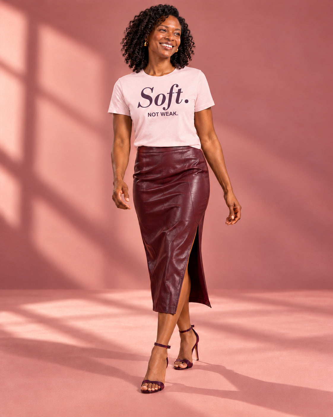 Amara wearing the Soft. Not Weak. affirmation tee in Soft Pink with a burgundy midi skirt and heels, full-length walking studio portrait