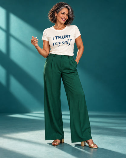 Freya wears the I Trust Myself affirmation tee in Natural with emerald trousers and gold heels, laughing in a teal studio.