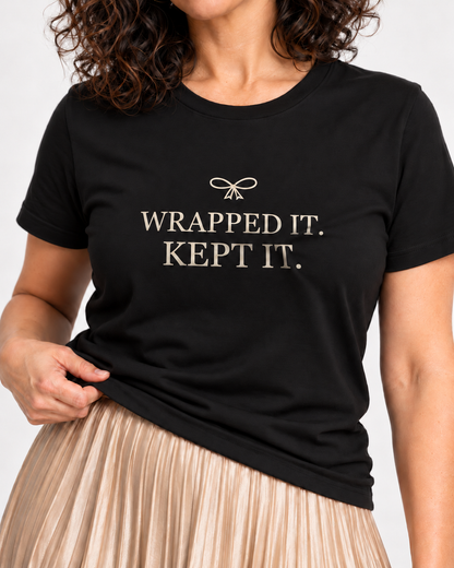 Close-up of cream Wrapped It. Kept It. serif lettering and a small ribbon bow on the Black crewneck T-shirt.