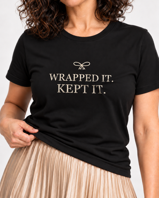 Close-up of cream Wrapped It. Kept It. serif lettering and a small ribbon bow on the Black crewneck T-shirt.