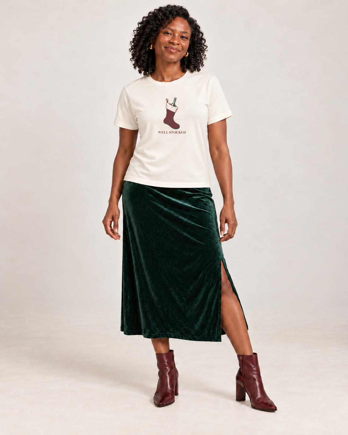 Amara wearing the Well Stocked Christmas graphic on a Natural Bella+Canvas 6400 women’s relaxed-fit tee, green velvet midi skirt and burgundy boots.